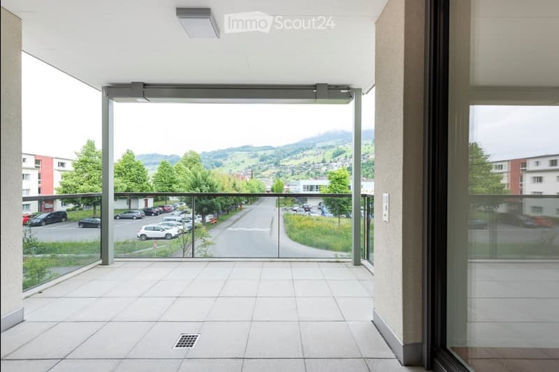 Modern balcony with large glass panels, tiled floor, and a scenic view of a town and surrounding hills.