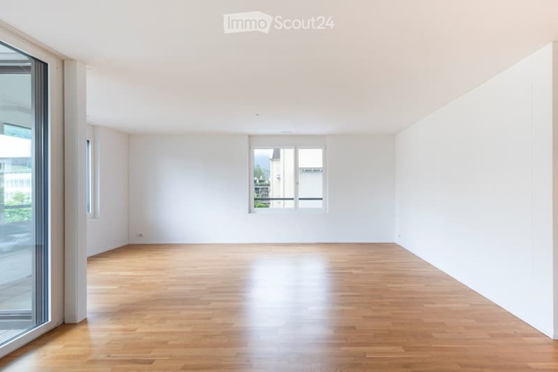 Empty room with light wooden flooring and white walls, featuring large windows and a sliding glass door.