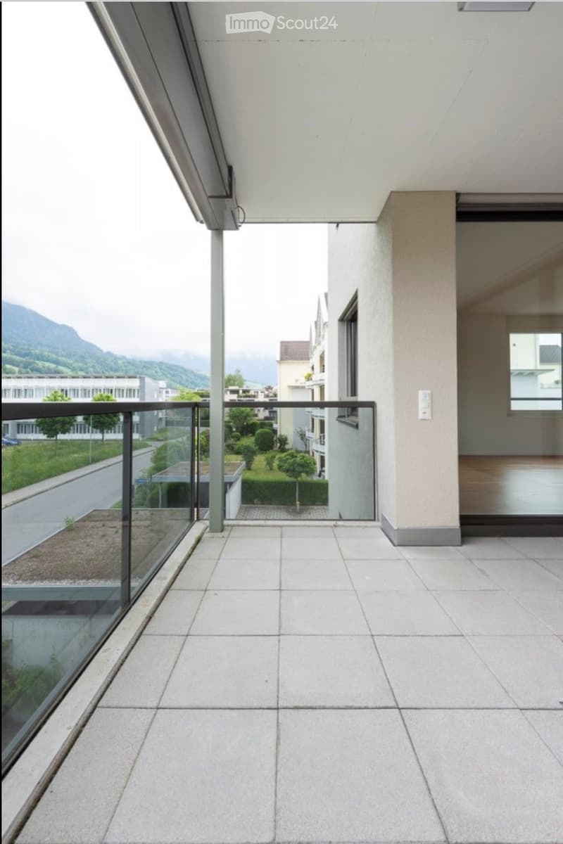 Modern balcony with white tiled flooring, glass railing, and a view of a landscaped area and distant mountains.