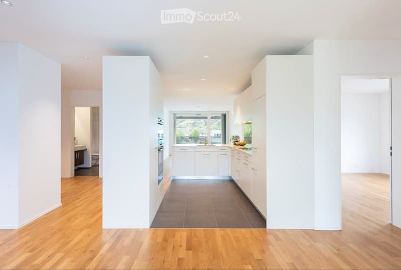 Modern, minimalist kitchen with white cabinets, a central island, and a large window overlooking a scenic view. The space features a combination of wooden flooring and tiled countertops.