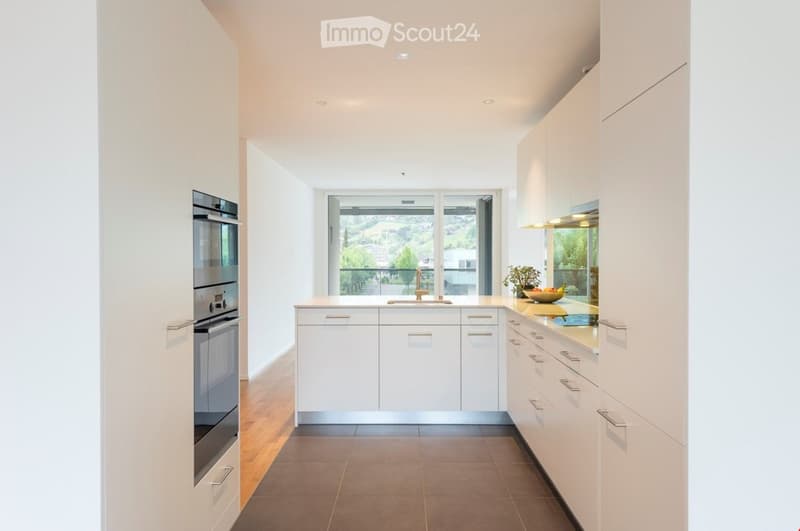 Modern kitchen with white cabinets, stainless steel oven, central island, and a view through large windows.