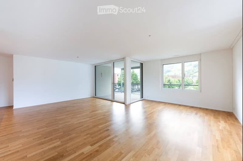 Empty room with wooden flooring, white walls, large windows, and sliding glass doors leading to a balcony.