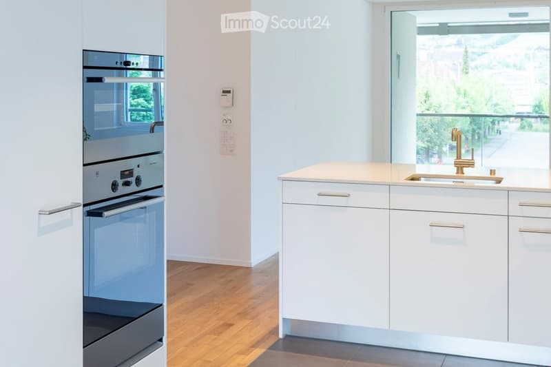 Modern kitchen with white cabinets, stainless steel oven, and a light-colored countertop with a gold faucet.