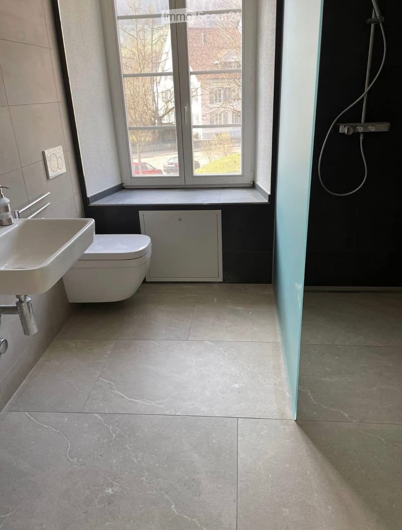 Modern bathroom with a sink, toilet, and shower area. The floor is tiled in a light grey marble pattern, and the walls are tiled in a light beige color. A large window provides natural light and a view of the street outside.