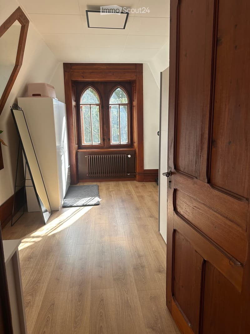 A narrow hallway with wooden flooring, arched windows at the end, and a radiator below them. A small rug is placed on the floor, and a mirror leans against the wall on the left side.
