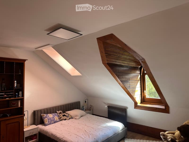 Cozy attic bedroom with slanted wooden ceiling, skylight windows, grey upholstered bed, nightstand with lamp, bookshelf, and stuffed animals on the bed and floor.