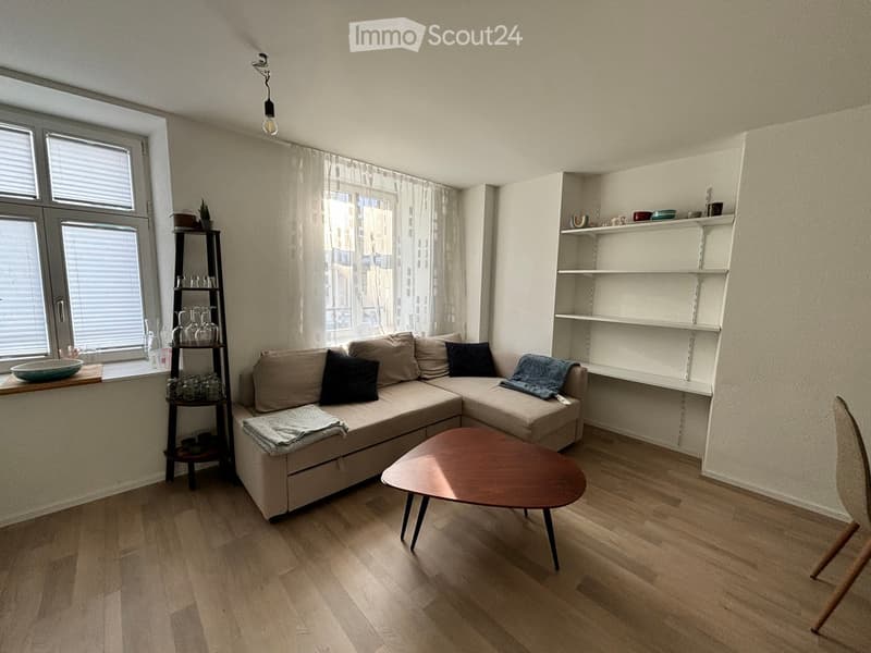 Modern living room with a sectional sofa, coffee table, shelves, and a view of the city through large windows.
