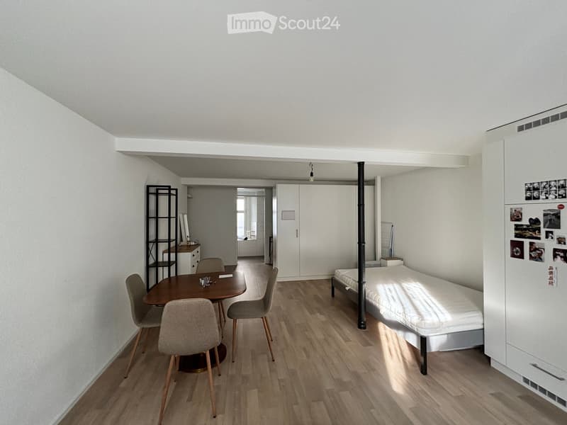A compact, modern studio apartment with a bed, dining table, chairs, a shelf, and a refrigerator. The room features light wooden flooring and white walls.