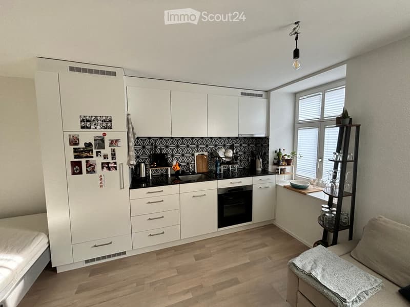 Modern kitchen and living area with white cabinets, black backsplash, wooden floor, and a small couch with a blanket.