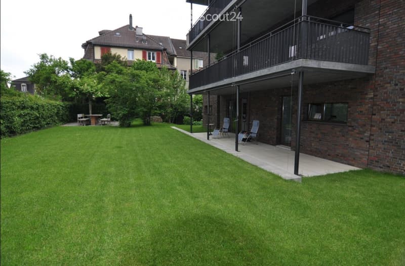 Well-maintained lawn with a small seating area, a modern building with a balcony, and additional outdoor furniture nearby.