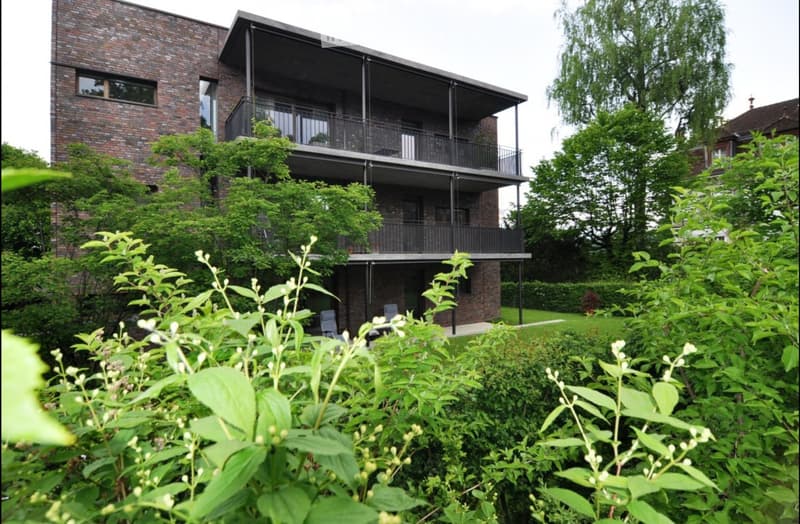 Modern brick building with multiple balconies and lush greenery in the foreground.