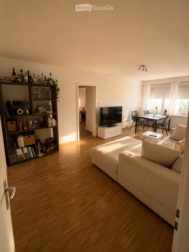 Bright living room with wooden flooring, a beige sofa, coffee table, bookshelf, dining table with chairs, and a television on a stand.