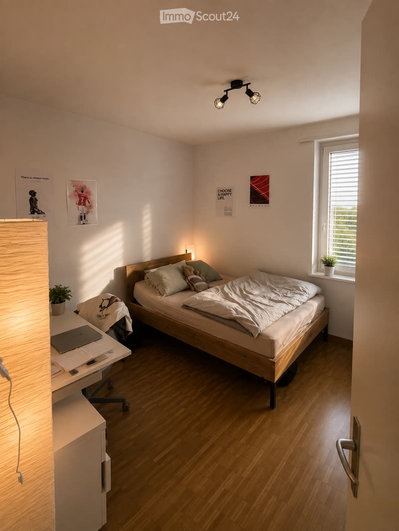 Cozy bedroom with a wooden bed, white bedding, a desk with a laptop, a chair, and a small plant on the windowsill. The room features wooden flooring and several posters on the walls.