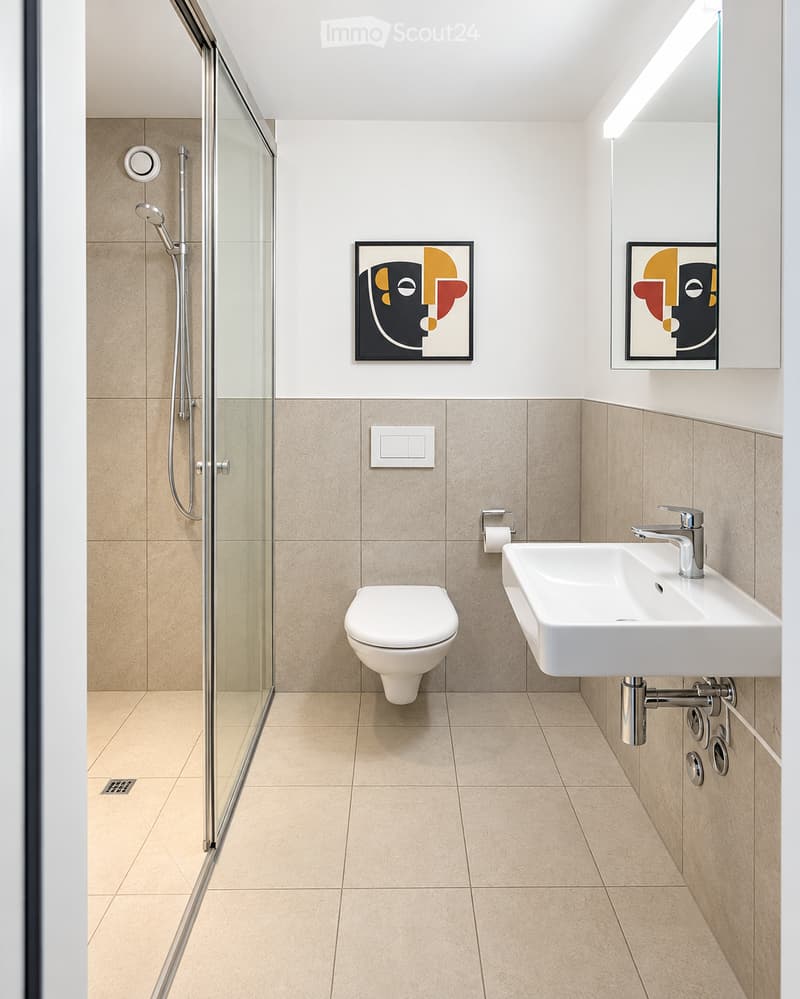 Modern bathroom with beige tiles, white toilet, sink, and glass shower enclosure. Features two abstract art pieces on the walls and a built-in mirror with lighting.