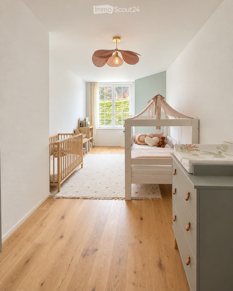 Bright, airy bedroom with wooden flooring, a crib, a bed with a canopy, a bookshelf, and a cabinet.