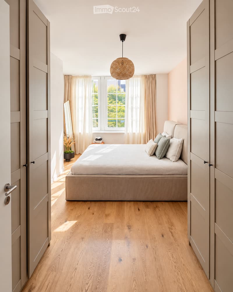 Modern bedroom with light wooden flooring, a large bed with pillows, a nightstand with a mirror, and a hanging woven light fixture.
