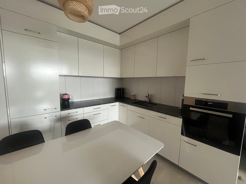 Modern kitchen with white cabinets, black countertops, a built-in oven, a sink with a faucet, a coffee maker, and a small dining area with a table and chairs.