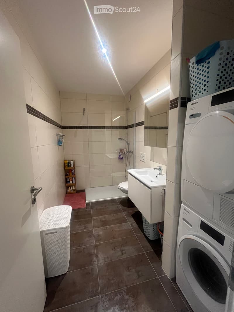 Modern bathroom with a white sink, toilet, shower, mirror, and a washing machine. Includes a laundry basket and a small shelf with toiletries.