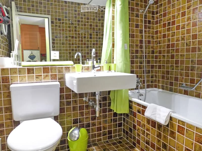 Bathroom with a white toilet, sink, and bathtub, surrounded by brown and beige tiled walls and floor. A green towel hangs on the side of the bathtub, and a mirror is mounted above the sink.