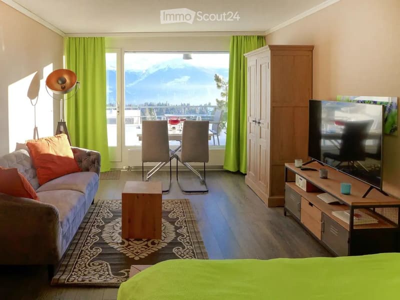 Bright living space with a sofa, coffee table, dining area with chairs and table, TV stand, wardrobe, and a scenic mountain view through large windows.