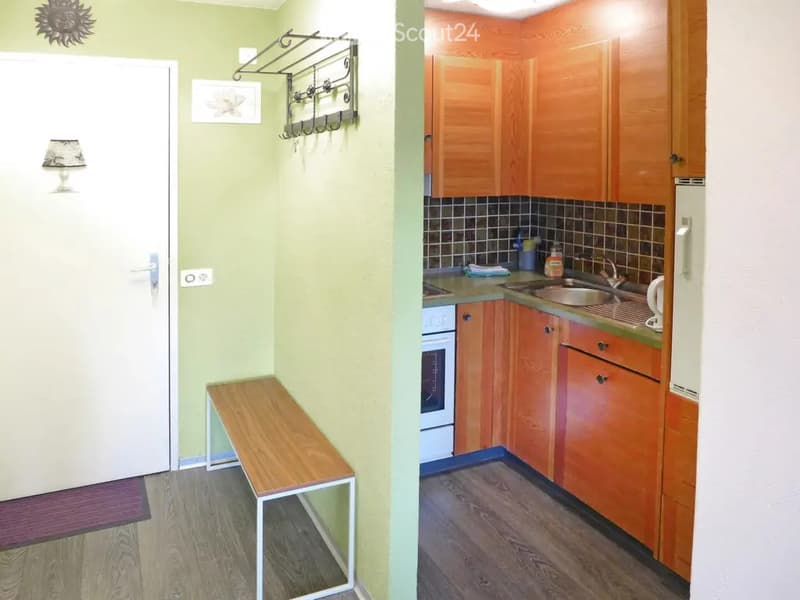 The image shows a small, modern kitchen with wooden cabinets, a sink, and a refrigerator. Adjacent to the kitchen is a small hallway with a wooden bench and a coat rack.
