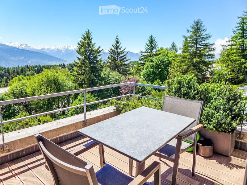 Balcony with a table and two chairs, overlooking a scenic mountain view with trees and greenery.