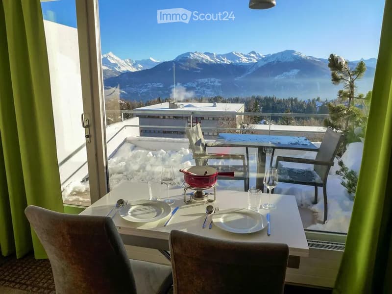 A cozy dining area with a table set for two, overlooking a snowy mountain landscape. The table is set with plates, cutlery, wine glasses, and a small pot on a candle holder. There are chairs around the table, and the room has large windows with green curt