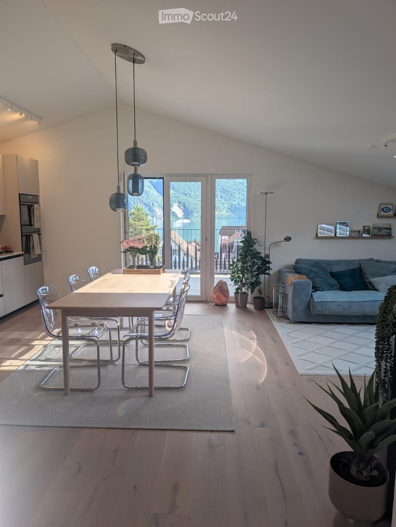 Bright, modern living and dining area with wooden floors, large windows offering a scenic view, a sofa, dining table with chairs, pendant lights, and decorative plants.