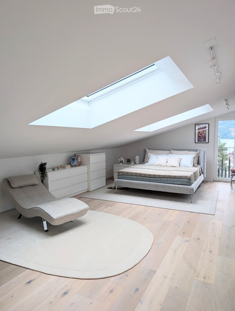 Modern bedroom with wooden flooring, large skylight windows, a grey bed with white pillows, a white dresser, a lounge chair, and a round rug.