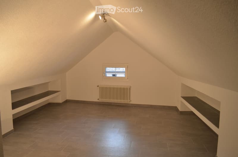 Empty attic room with slanted ceilings, tiled floor, built-in shelves, radiator, and a small window.