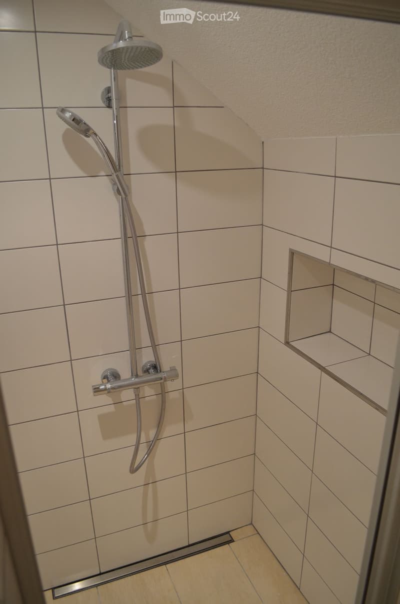 Modern shower area with beige tiled walls and floor, featuring a handheld showerhead and control handle.