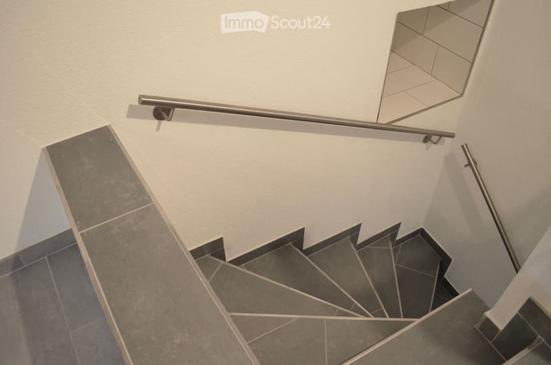 Modern staircase with grey tiled steps and a sleek metal handrail, set against white walls and a light-colored ceiling.