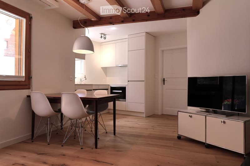 Open-plan living and dining area with wooden flooring, white chairs around a dark wooden table, a television on a cabinet, and a modern kitchen with white cabinets and an oven.