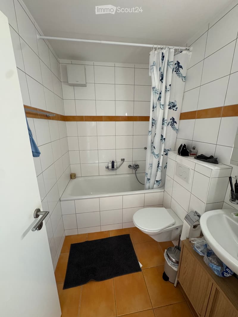 Bathroom with white tiled walls, a bathtub, toilet, sink, shower curtain, and a small wooden cabinet. The floor is tiled in a warm tone with a black bath mat placed in front of the bathtub.