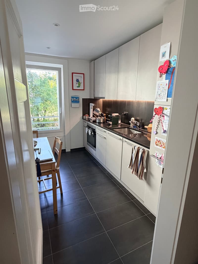 Modern kitchen with white cabinets, stainless steel appliances, dark tiled floor, and a small dining area with a table and chairs. A large window offers a view of greenery outside.