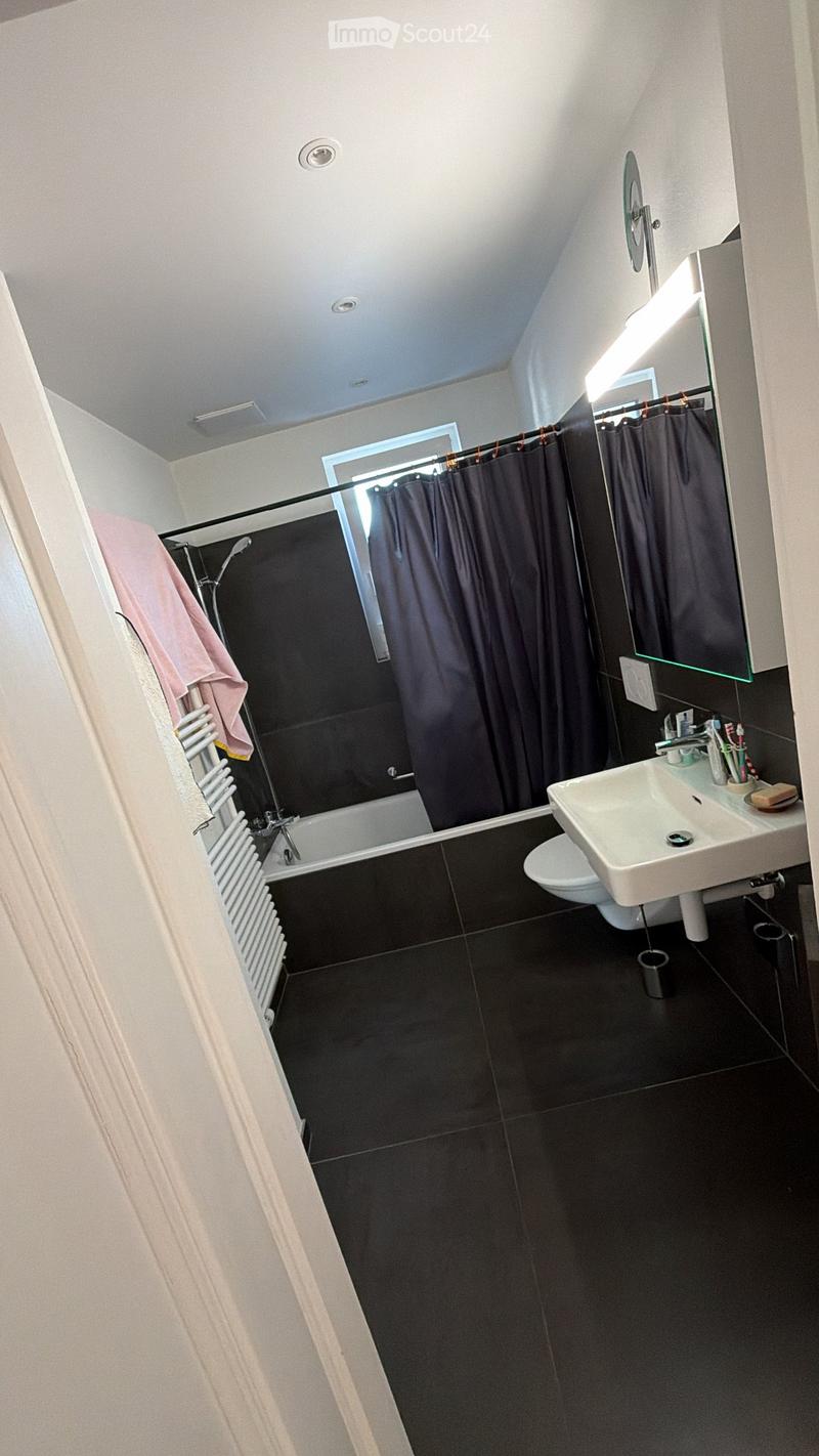 Modern bathroom with black tiles, bathtub, sink, toilet, mirror, and shower curtain. Includes a towel on a rack and a toothbrush holder on the sink.
