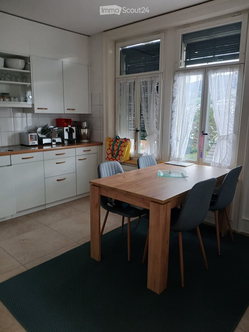 Bright kitchen with white cabinets, wooden dining table, four chairs, and various appliances on the counter.