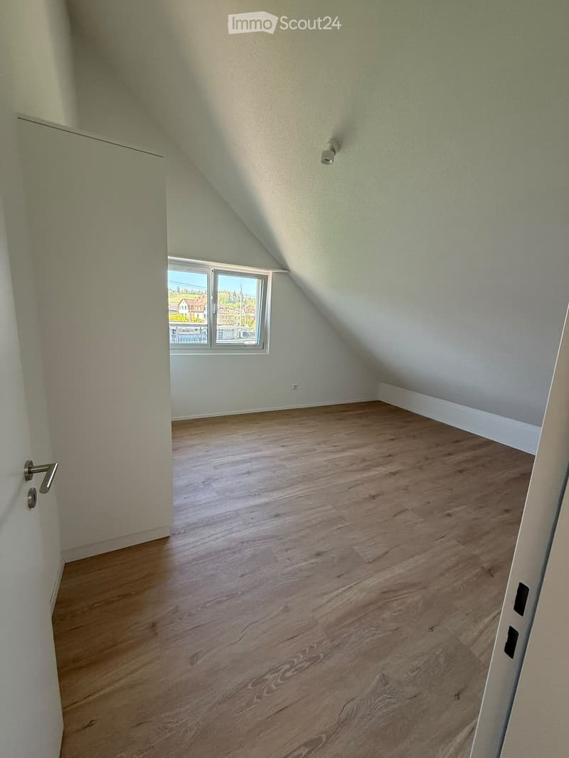 Empty attic room with wooden flooring, slanted ceilings, and a single window offering a view of the outside.