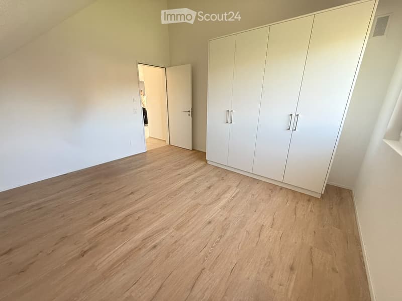 Empty room with light wooden flooring, white walls, and built-in white wardrobes. A doorway leads to another room.