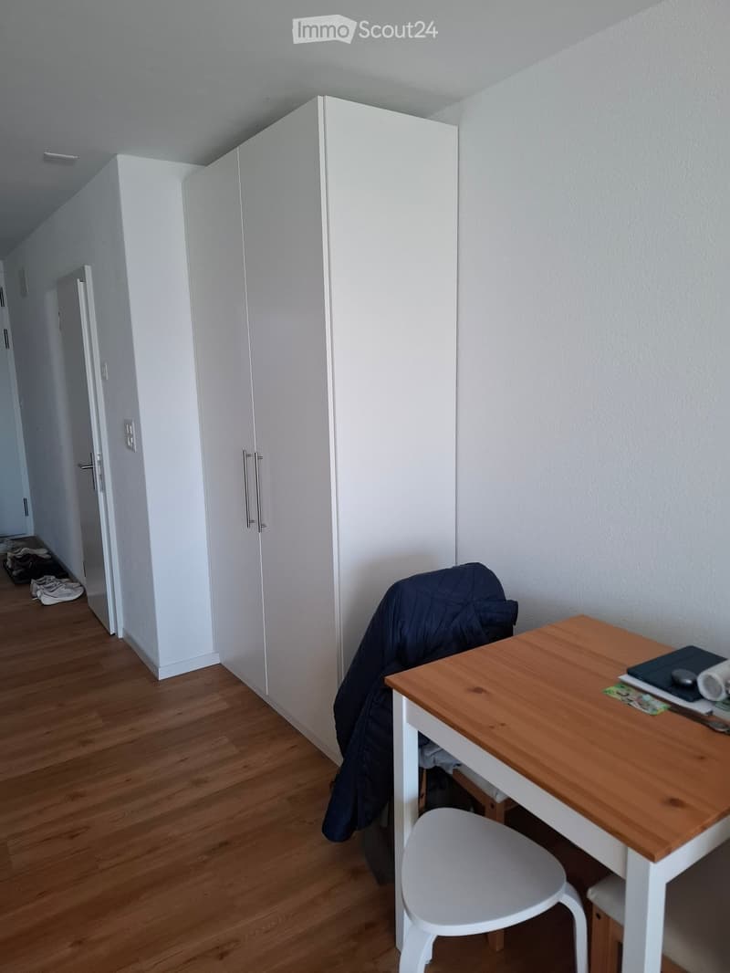 A small, furnished room with a wooden floor, a white wardrobe, a wooden table with a white chair, and a jacket draped over the chair.