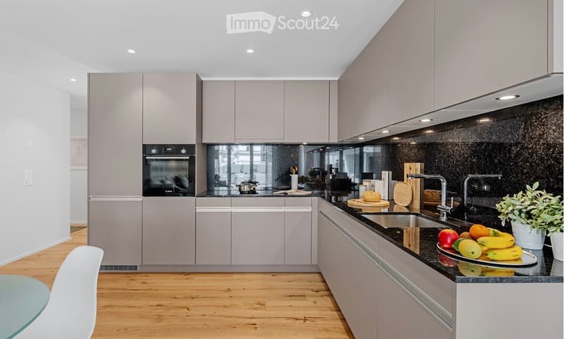 Modern kitchen with light grey cabinets, black countertops, stainless steel appliances, and a wooden floor. Features include an oven, sink, faucet, cutting board, bowl, and a small table with a chair.