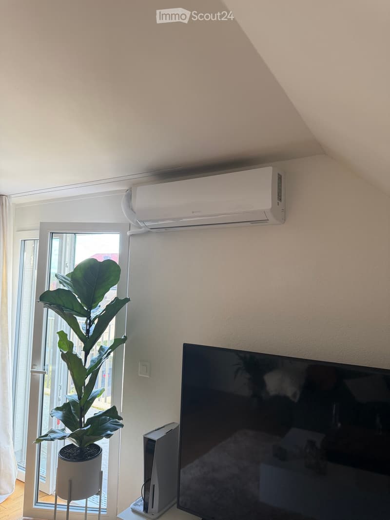 A modern living room with a large window, a potted plant, a television, and an air conditioner mounted on the wall.