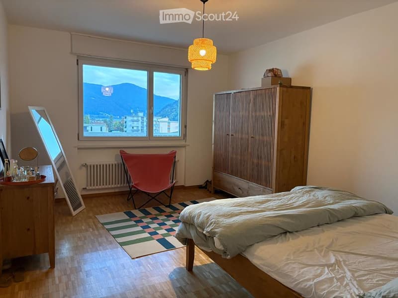 Cozy bedroom with wooden flooring, large window offering a mountain view, a neatly made bed, a wardrobe, a red folding chair, a small table with a mirror, and a hanging lamp.