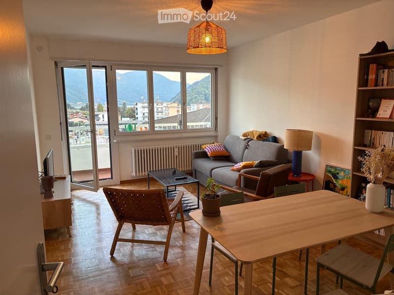 Modern living room with large windows offering a scenic mountain view, featuring a grey sofa, coffee table, wooden dining table, wicker chair, and decorative lamps.