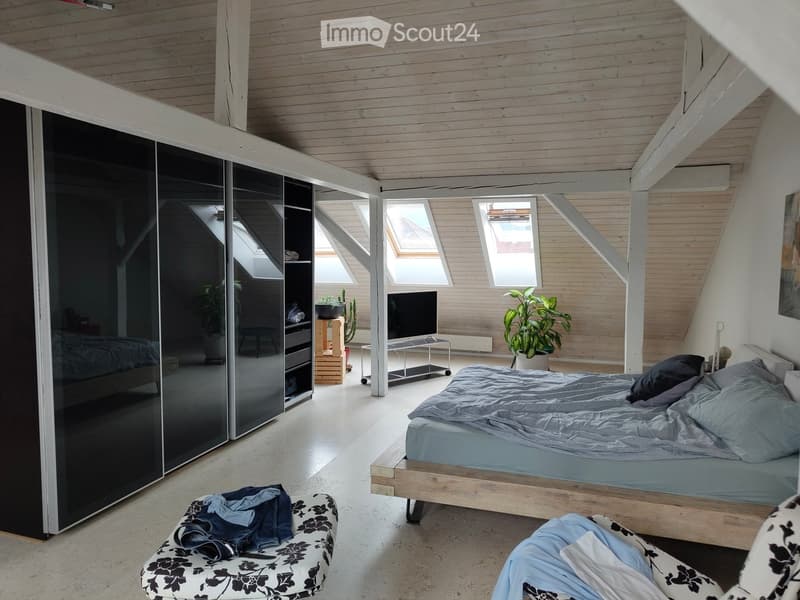 Modern bedroom with wooden bed, grey bedding, black and white patterned chairs, small table, potted plants, and a large mirrored wardrobe.