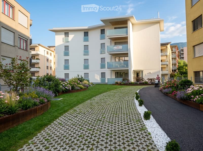 Well-maintained garden area with a paved pathway, green lawn, and various flowering plants and shrubs. A modern multi-story building with balconies is visible in the background.