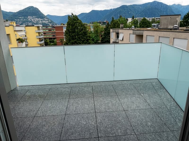 Modern balcony with grey tiled flooring and glass railing, overlooking a cityscape with mountains in the background.