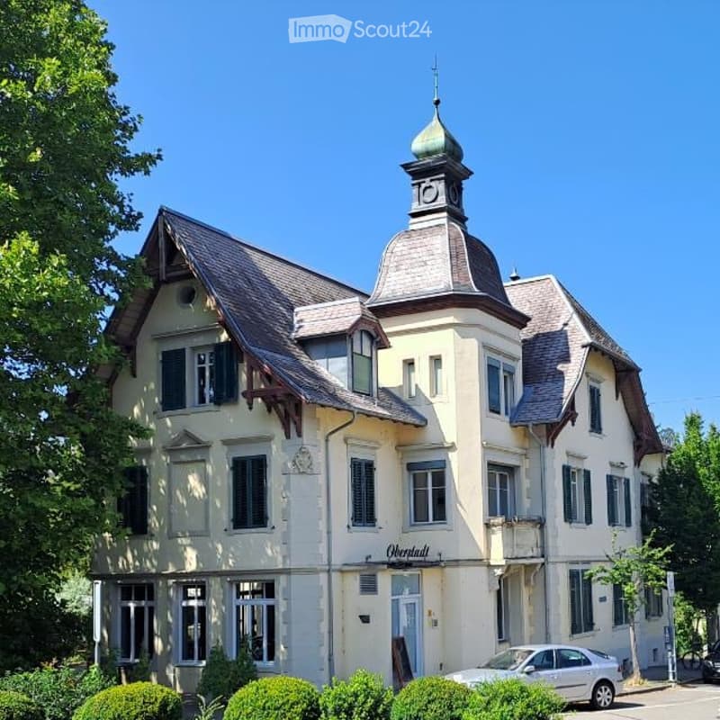 A two-story building with a distinctive dome-shaped roof and clock tower, surrounded by greenery and a parked car.