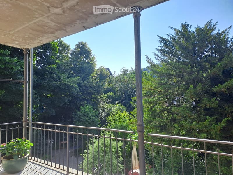 Balcony with metal railing, potted plant, and umbrella, overlooking a lush green garden area.