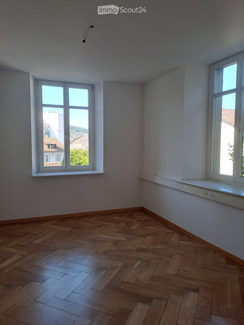 Empty room with wooden herringbone flooring, white walls, and large windows offering a view of neighboring buildings and greenery.